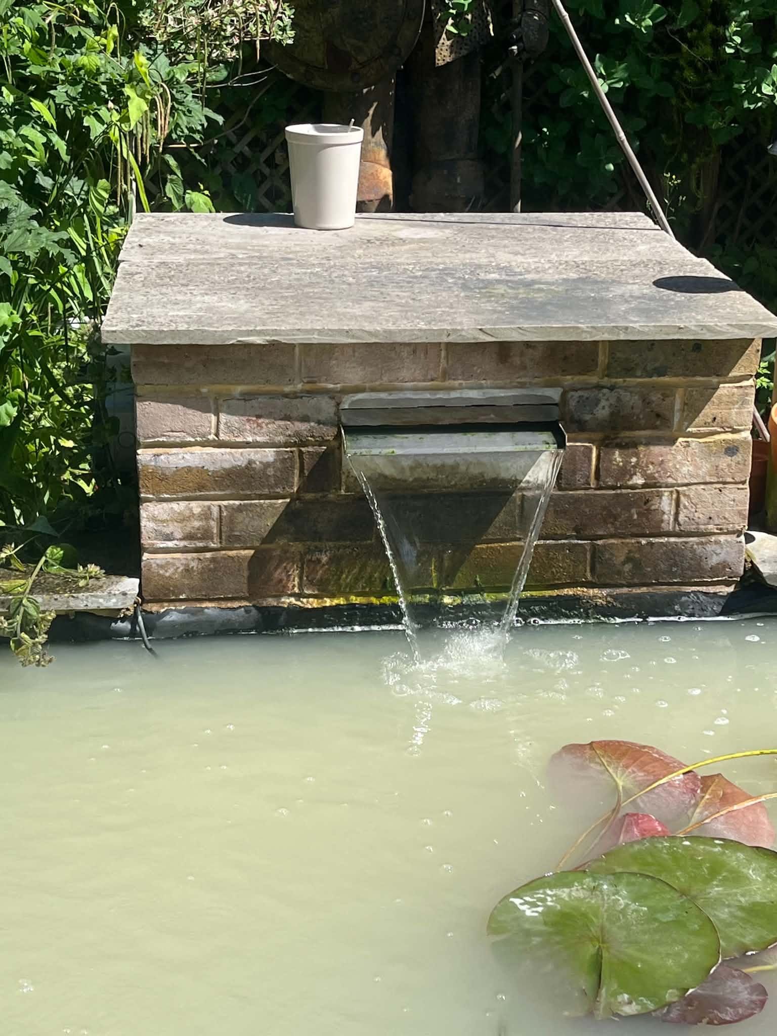 A brick water feature with a stone cap, with water falling from a metal spout into the pond below